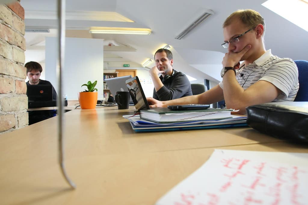 People working at a long wooden table in a bright attic coworking space with a brick wall, skylights, and a potted plant.