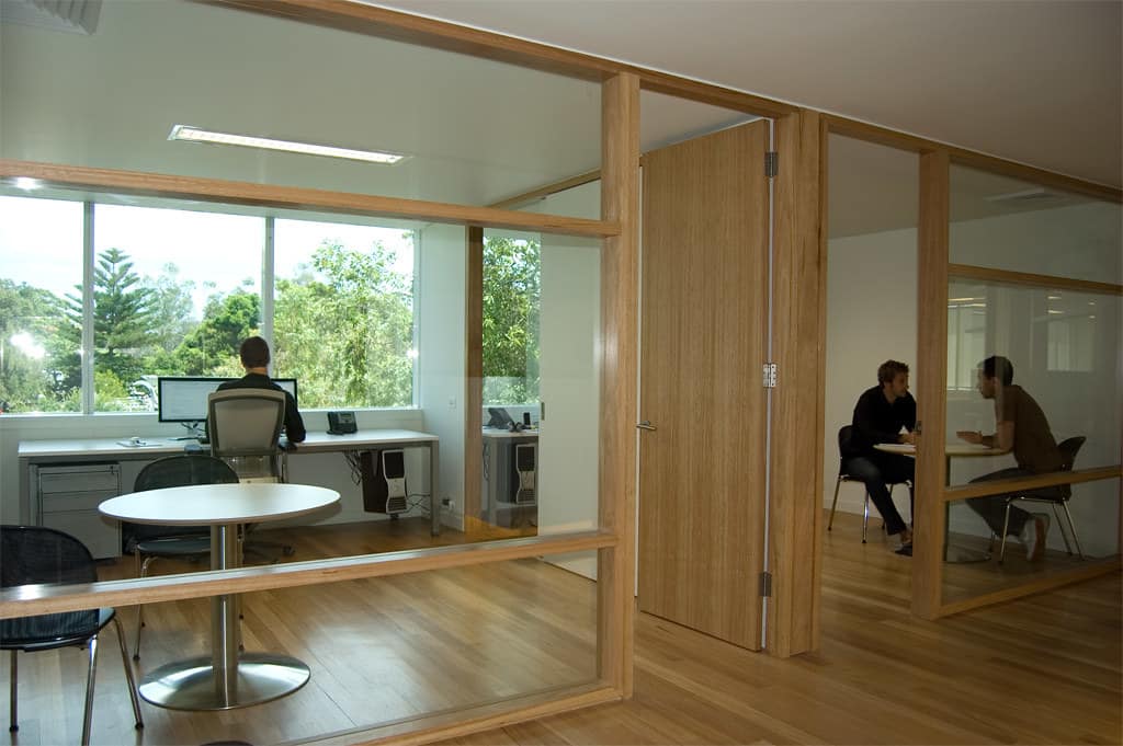 Glass-walled private offices framed in light wood, with one person working at a desk by a tree-lined window and two people talking at a table in the next room.